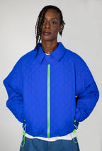 A model wearing a bright cobalt blue quilted jacket with a neon green zipper and matching drawstrings, paired with a white undershirt and blue jeans, against a plain light gray background.