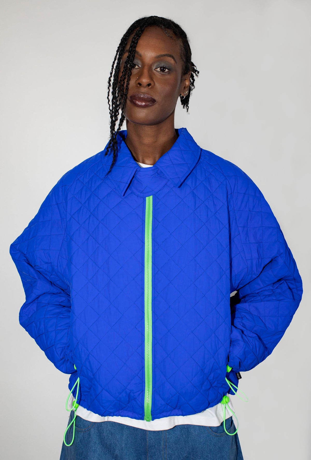 A model wearing a bright cobalt blue quilted jacket with a neon green zipper and matching drawstrings, paired with a white undershirt and blue jeans, against a plain light gray background.