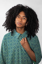 A person with voluminous curly black hair wearing a green and blue patterned button-up shirt, photographed against a plain light gray background.