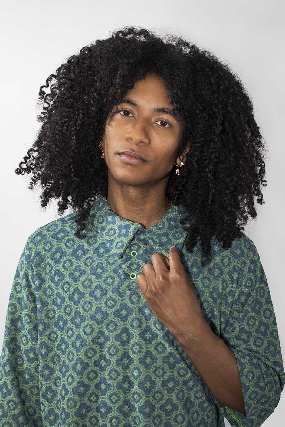 A person with voluminous curly black hair wearing a green and blue patterned button-up shirt, photographed against a plain light gray background.