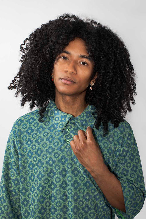 A person with voluminous curly black hair wearing a green and blue patterned button-up shirt, photographed against a plain light gray background.