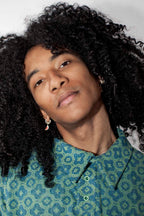 A close-up portrait of a person with voluminous dark curly hair, wearing a blue and green patterned button-up shirt and small hoop earrings, against a plain light background.