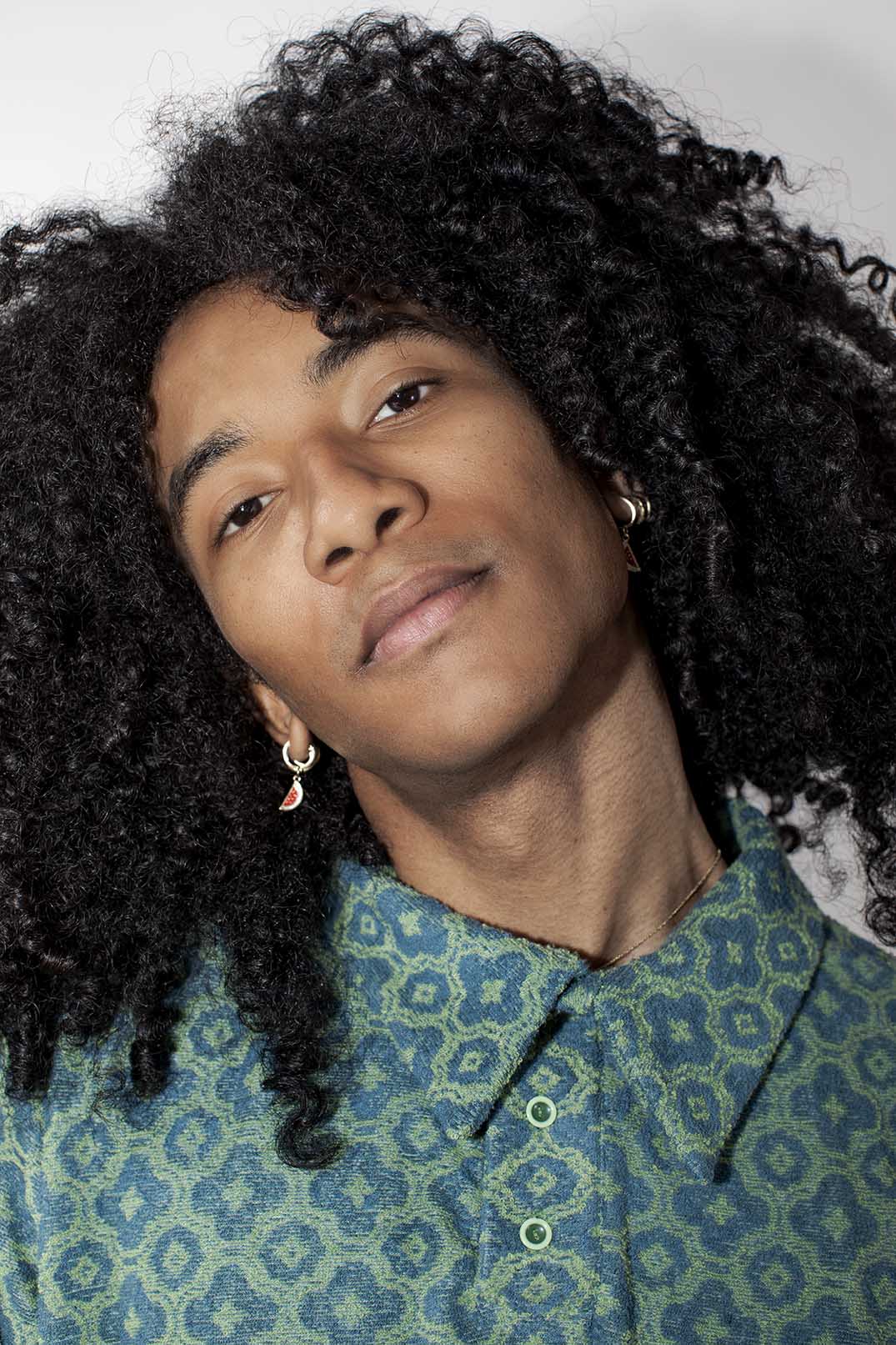 A close-up portrait of a person with voluminous dark curly hair, wearing a blue and green patterned button-up shirt and small hoop earrings, against a plain light background.