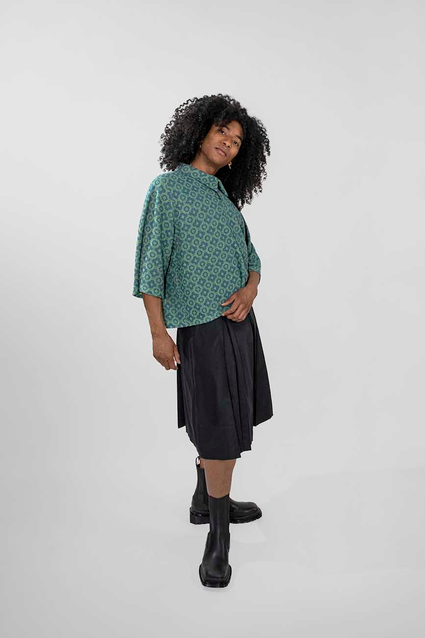 A person modeling a green patterned short-sleeve polo top and a dark pleated skort with black boots, standing against a light gray studio background.