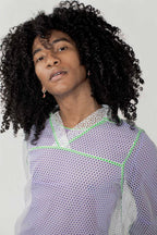 A person with voluminous curly black hair wearing a white mesh long-sleeve top with lime green geometric trim, photographed against a plain light gray background.