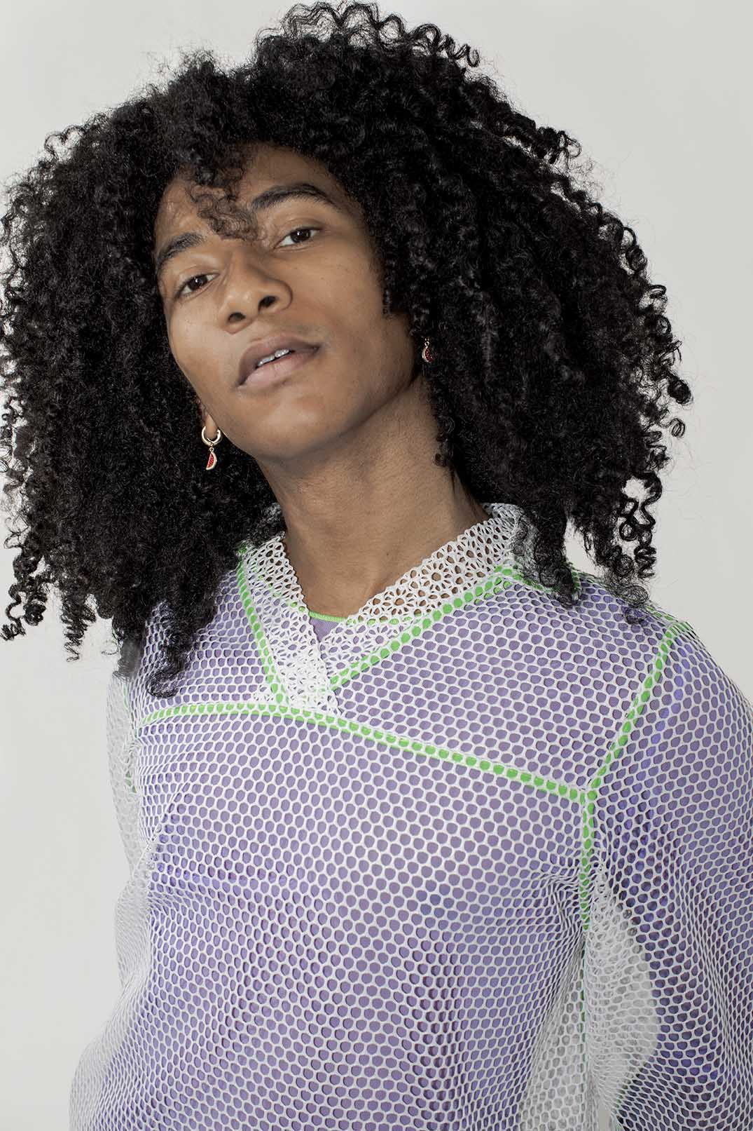 A person with voluminous curly black hair wearing a white mesh long-sleeve top with lime green geometric trim, photographed against a plain light gray background.