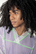 A close-up portrait of a person with voluminous dark curly hair wearing a lavender-purple mesh top with a white bigger mesh polo t-shirt and bright green geometric accents.