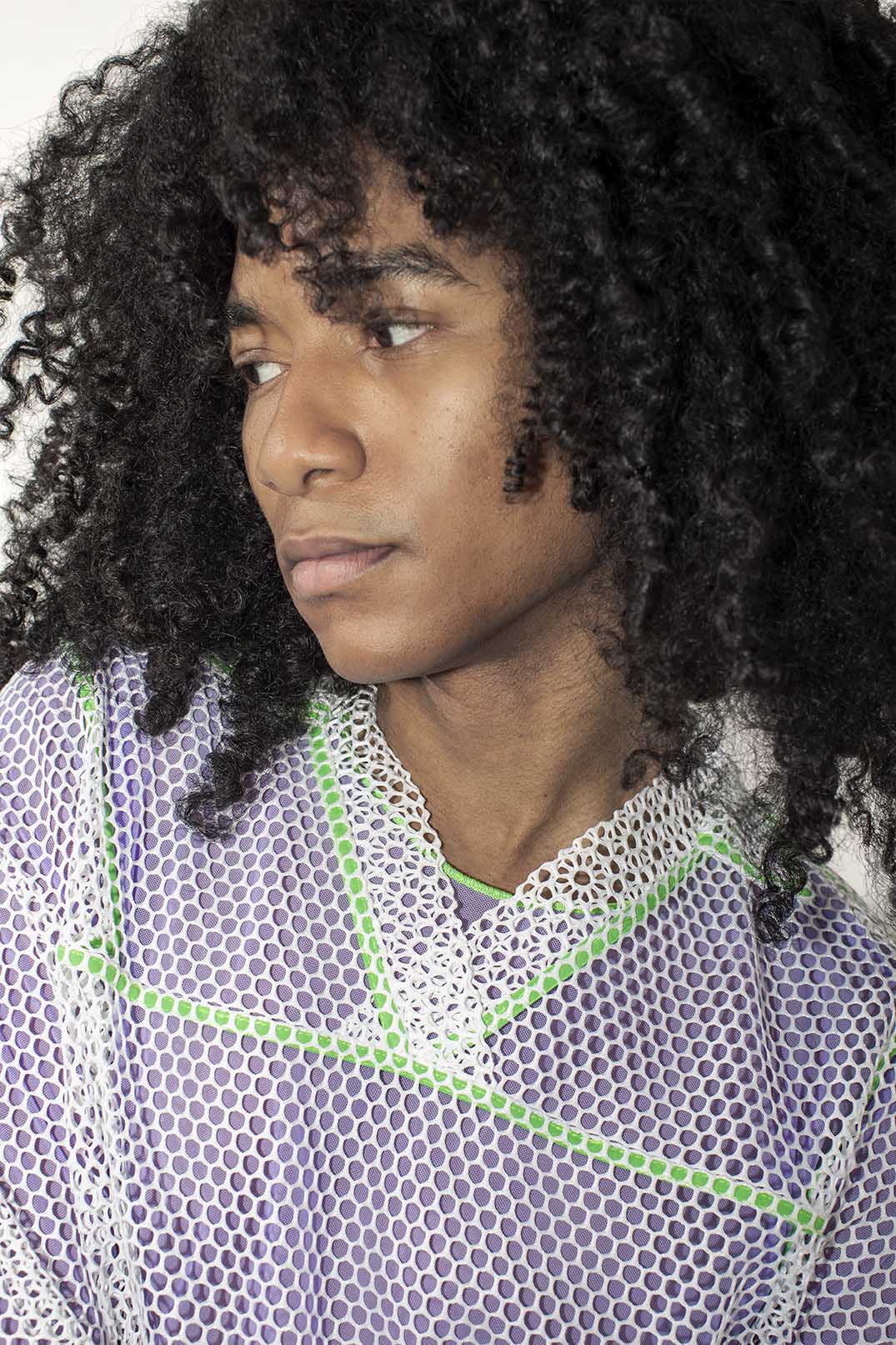 A close-up portrait of a person with voluminous dark curly hair wearing a lavender-purple mesh top with a white bigger mesh polo t-shirt and bright green geometric accents.