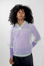 A model with curly black hair wearing a lavender and white mesh long-sleeve top with lime green trim, paired with black pants, against a light gray background.