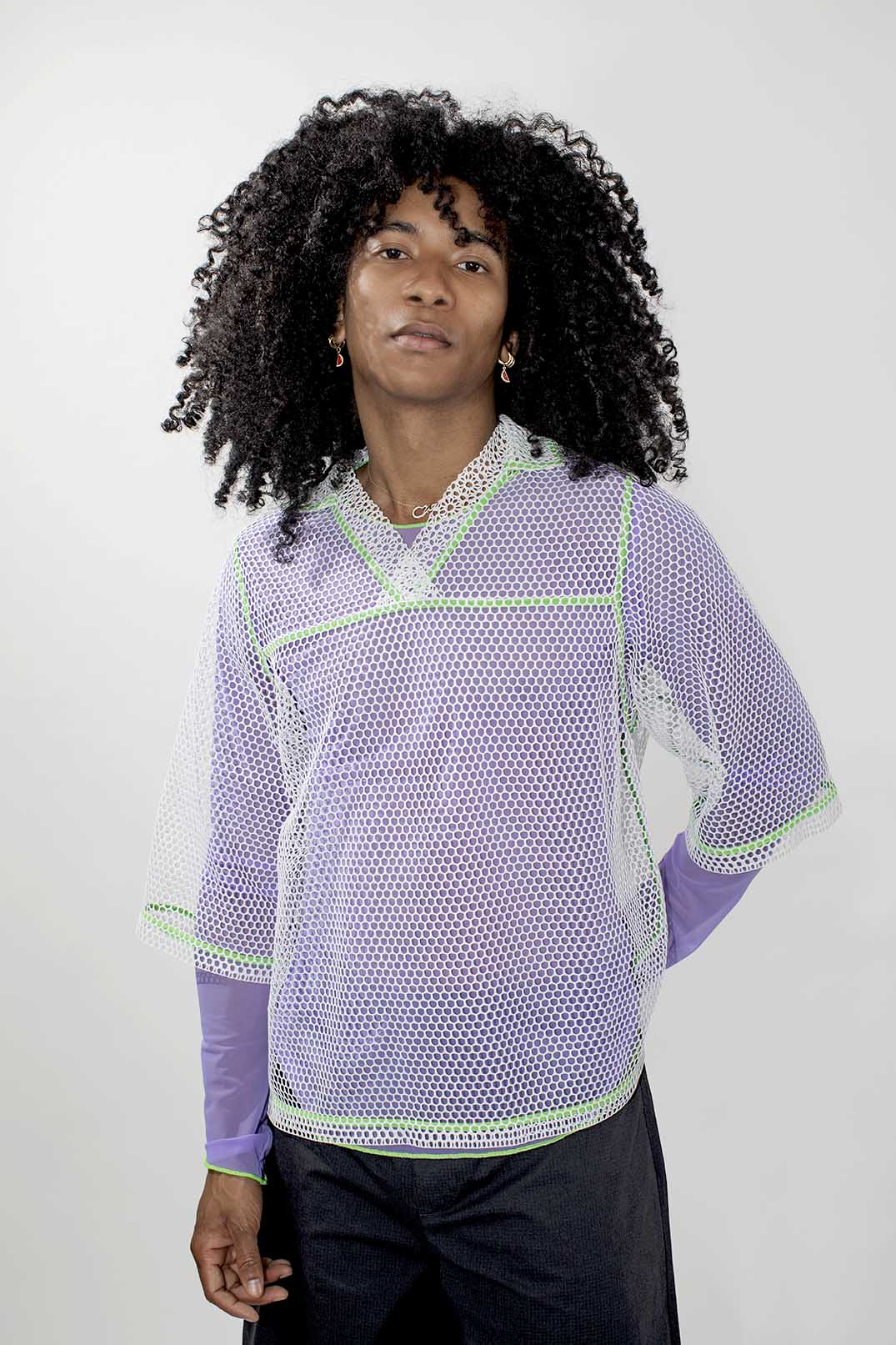 A model with curly black hair wearing a lavender and white mesh long-sleeve top with lime green trim, paired with black pants, against a light gray background.