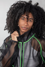 A person with voluminous curly black hair wearing a black sheer mesh zip jacket with bright neon green piping, photographed against a light gray studio background.