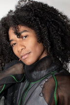 A close-up portrait of a person with voluminous curly black hair wearing a black sheer mesh jacket with green piping, against a plain light background.