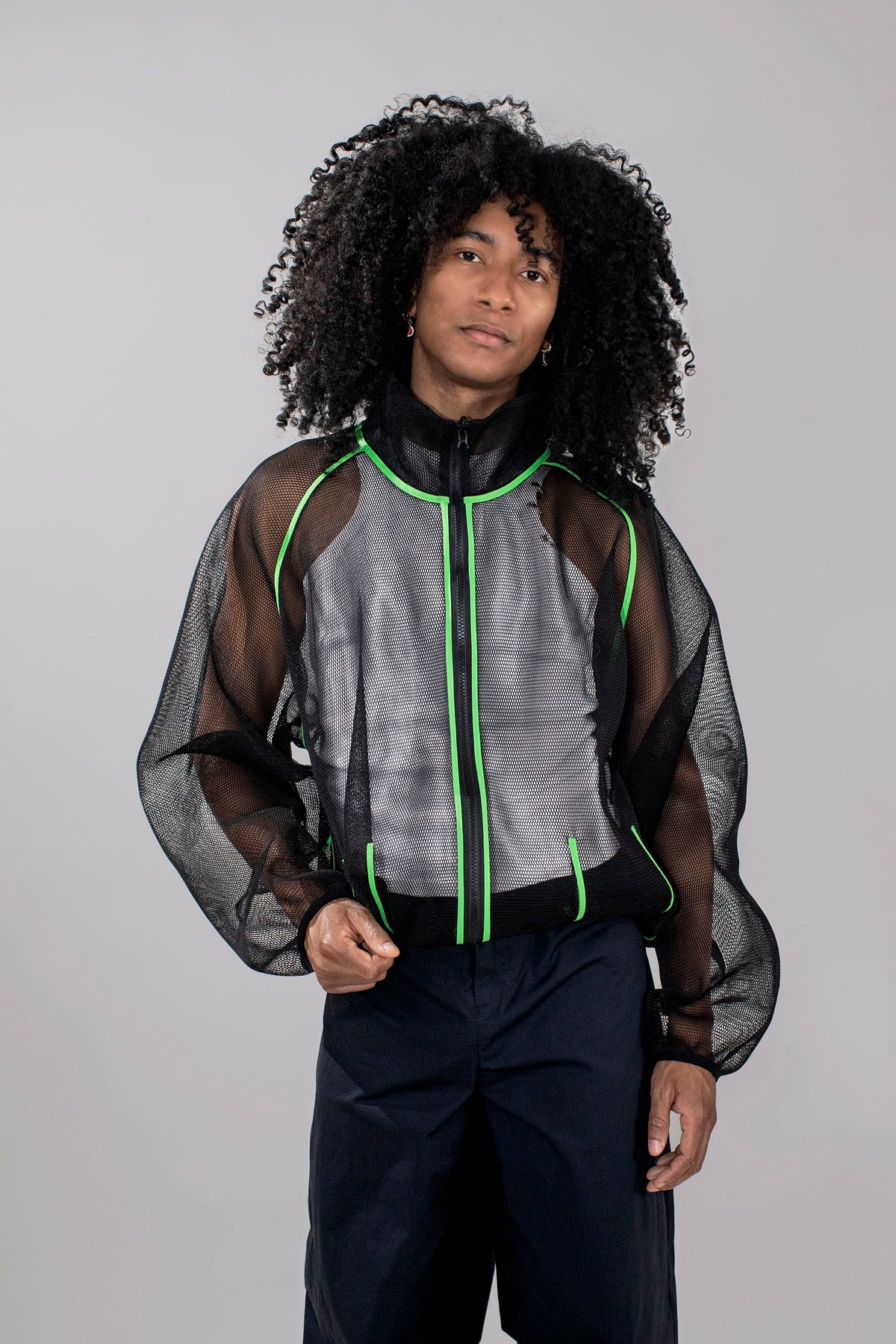 A model with curly black hair wearing a black mesh jacket with bright green piping, paired with dark trousers, against a solid light gray background.