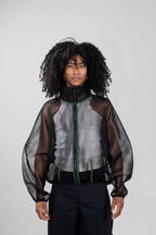 A model with curly black hair wearing a black sheer mesh zip-up jacket over a white top and black pants, photographed against a light gray studio background.