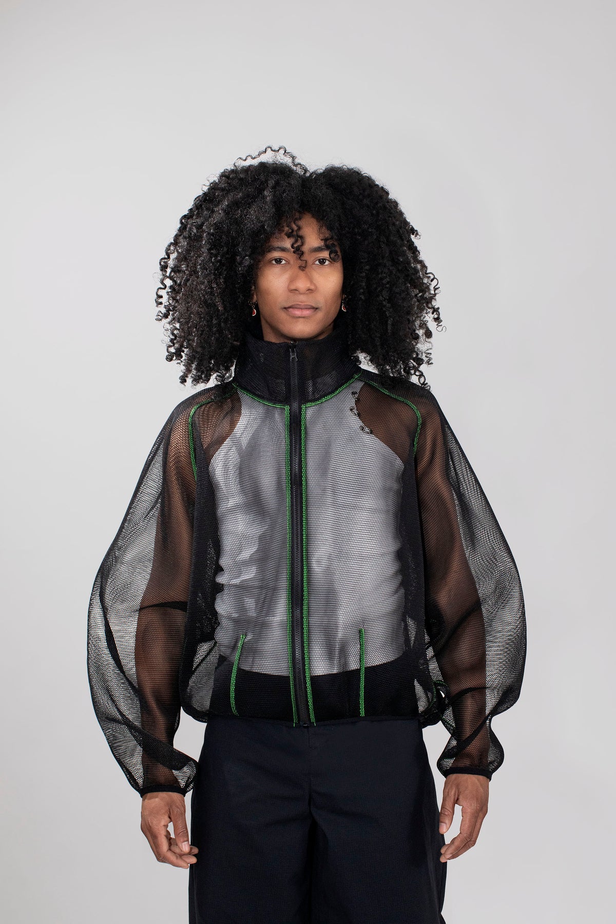 A model with curly black hair wearing a black sheer mesh zip-up jacket over a white top and black pants, photographed against a light gray studio background.