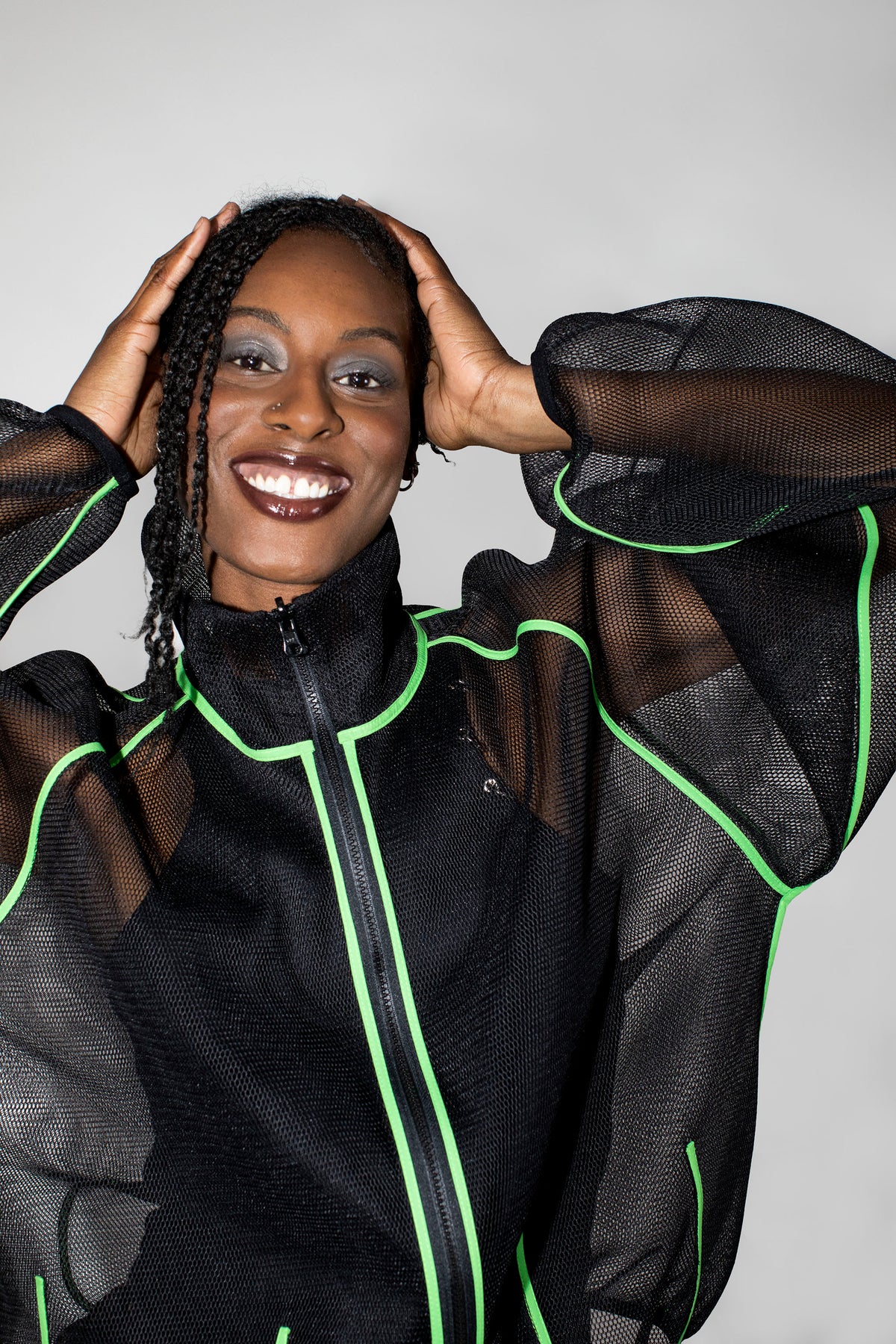 A smiling woman with braided hair poses in a black sheer mesh zip-up jacket with bright neon green piping details, set against a plain light gray background.