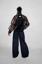 A model shown from the back wearing a black mesh jacket with neon green piping, paired with wide-leg black trousers, against a light gray studio background.