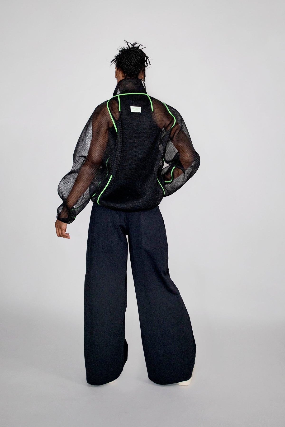 A model shown from the back wearing a black mesh jacket with neon green piping, paired with wide-leg black trousers, against a light gray studio background.