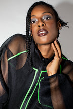A close-up portrait of a model wearing a black sheer mesh zip-up jacket with bright neon green piping details, set against a plain light gray background.