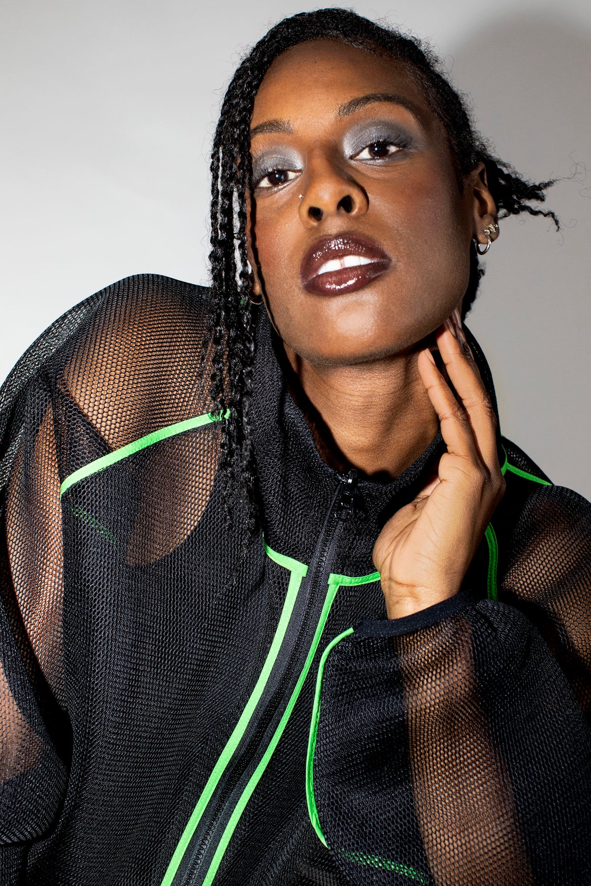 A close-up portrait of a model wearing a black sheer mesh zip-up jacket with bright neon green piping details, set against a plain light gray background.