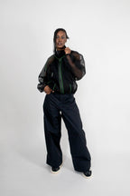 A model stands against a light gray studio background wearing a black sheer mesh zip-up jacket with green piping details, paired with wide-leg black trousers and black sneakers with white soles.