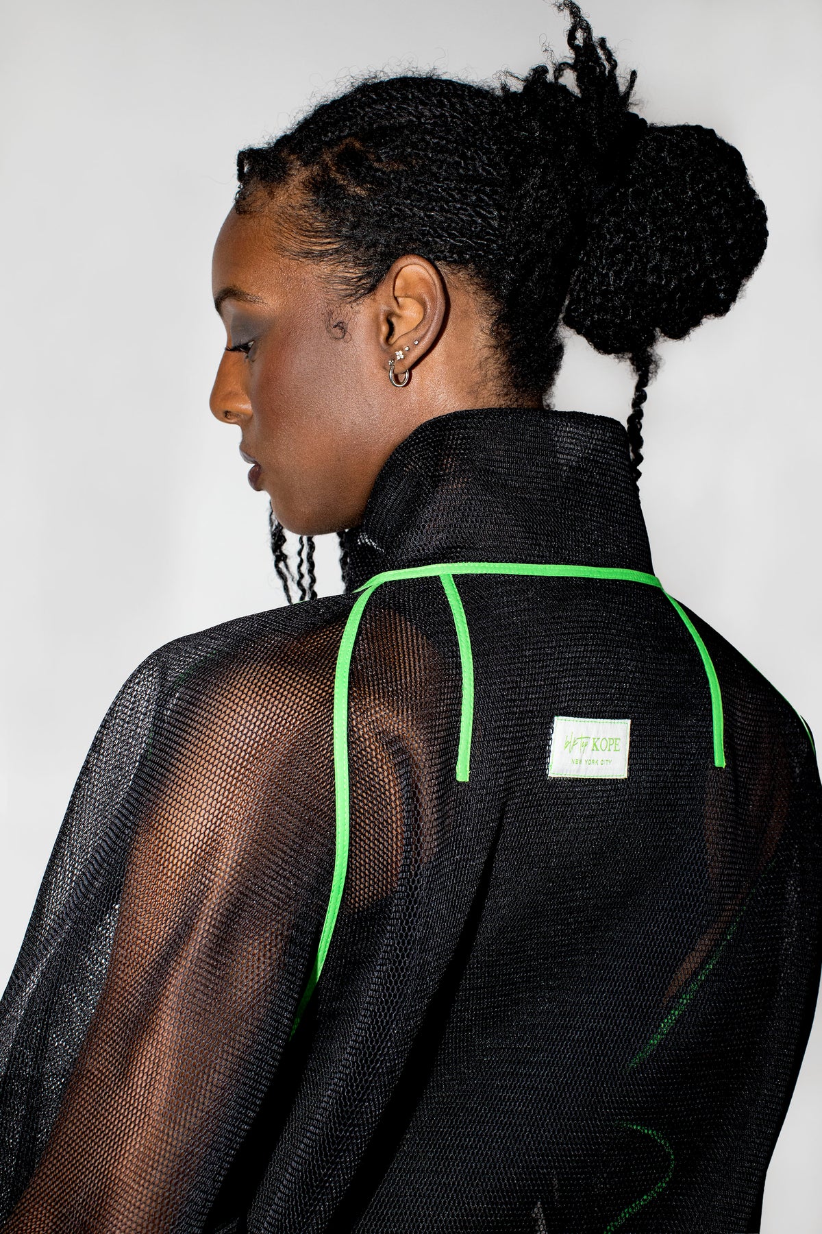 A model shown from the back wearing a black sheer mesh high-neck jacket with bright neon green piping details and a small white brand label.