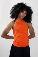 A model with voluminous curly black hair wearing a bright orange sleeveless ribbed tank top and black pants, standing against a light gray studio background.