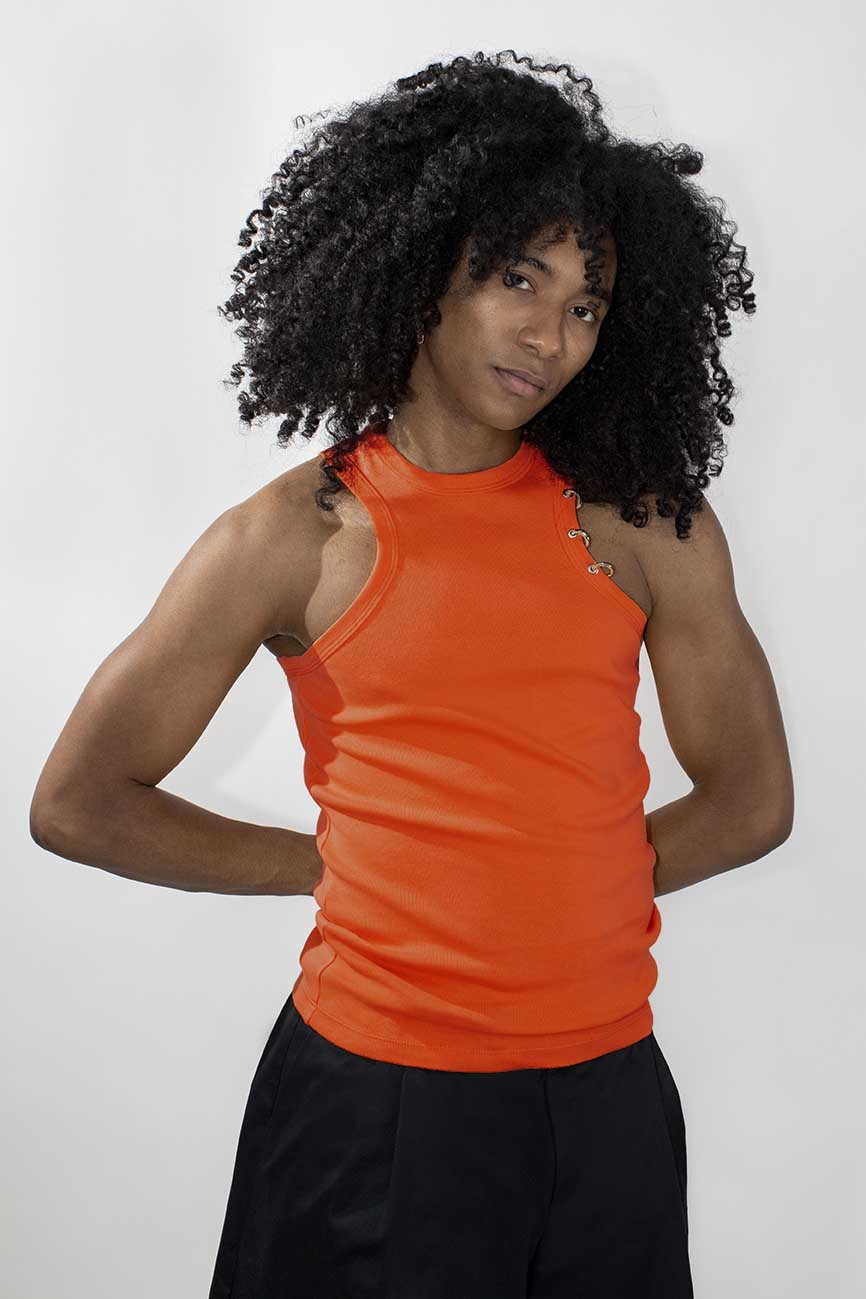 A model with voluminous curly black hair wearing a bright orange sleeveless ribbed tank top and black pants, standing against a light gray studio background.