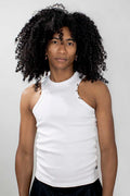 A person with voluminous curly black hair wearing a white sleeveless mock-neck top with small cutout details on the shoulders, paired with black bottoms, against a light gray studio background.