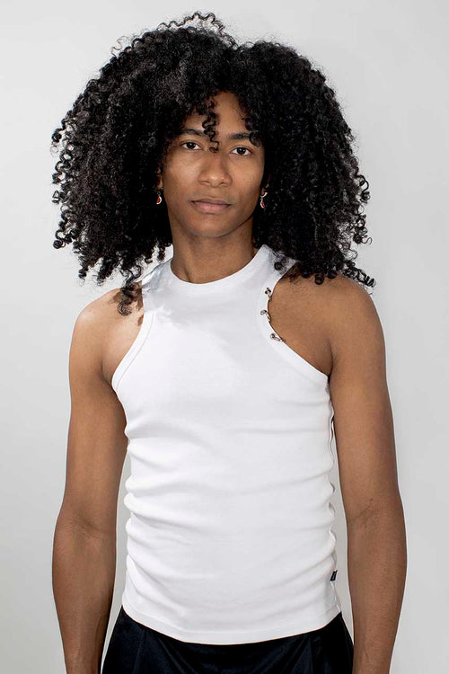 A person with voluminous curly black hair wearing a white sleeveless mock-neck top with small cutout details on the shoulders, paired with black bottoms, against a light gray studio background.