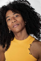 A close-up portrait of a person with voluminous curly black hair wearing a yellow sleeveless top and small silver hoop earrings against a plain light background.