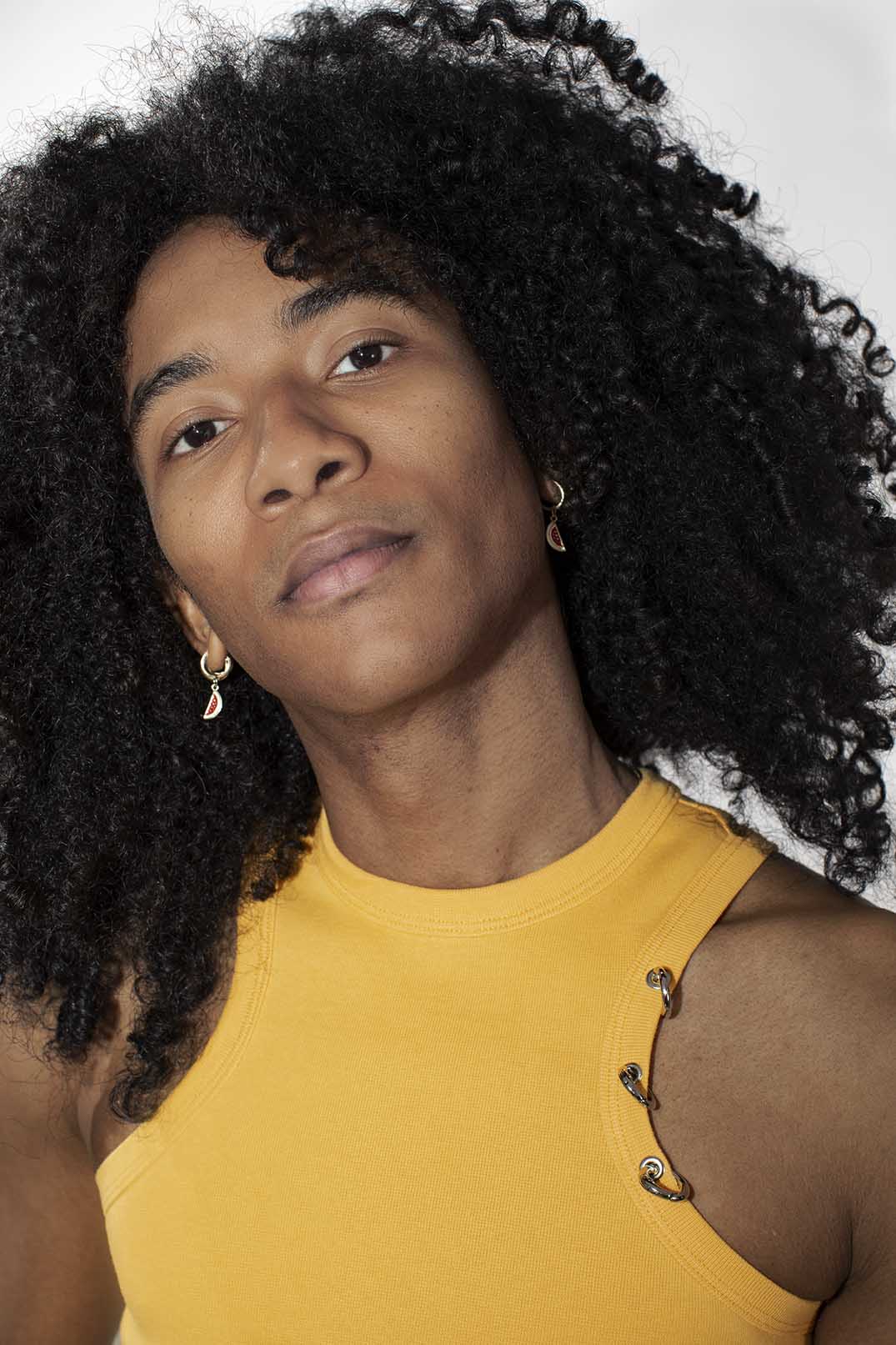 A close-up portrait of a person with voluminous curly black hair wearing a yellow sleeveless top and small silver hoop earrings against a plain light background.