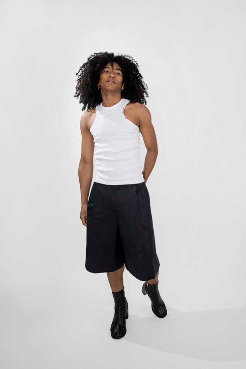 A model with curly black hair wearing a white sleeveless top with a cutout, black wide-leg shorts, and black boots, standing against a light gray studio background.