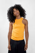 A person with voluminous curly black hair wearing a bright yellow sleeveless top and black pants, standing against a plain light gray studio background.
