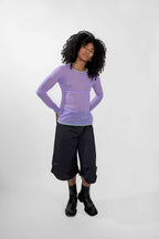 A model with curly black hair wearing a sheer light purple long-sleeve top, dark cropped pants, and black boots, standing against a light gray studio background.