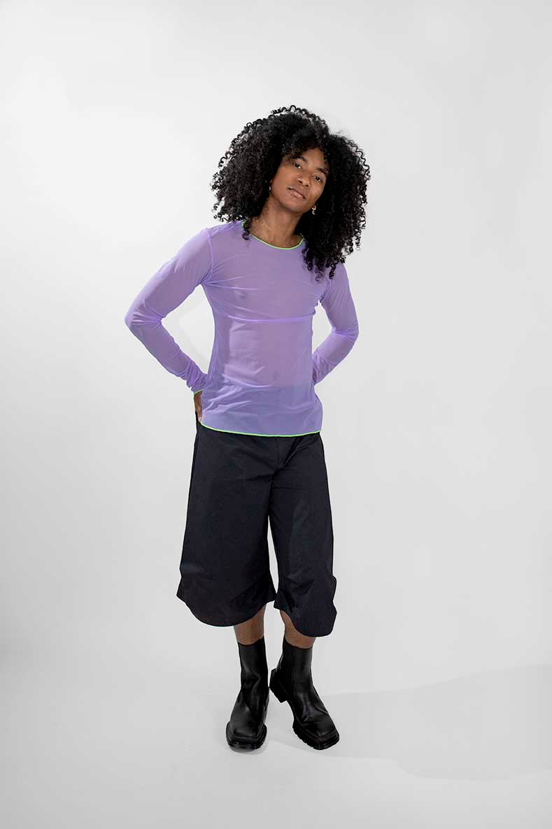 A model with curly black hair wearing a sheer light purple long-sleeve top, dark cropped pants, and black boots, standing against a light gray studio background.
