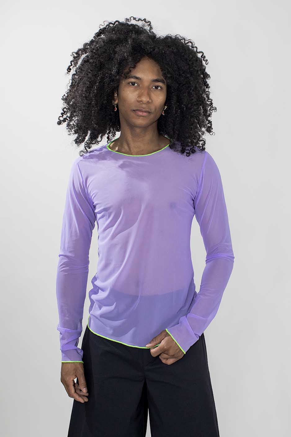 A model with curly black hair wearing a light purple sheer long-sleeve shirt with a thin lime green collar and cuffs, paired with black pants, against a light gray background.