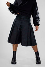 A model wearing a black pleated midi skort, a black long-sleeve top with white graphic prints on the sleeves, and black ankle boots, shown from the waist down against a light gray studio background.