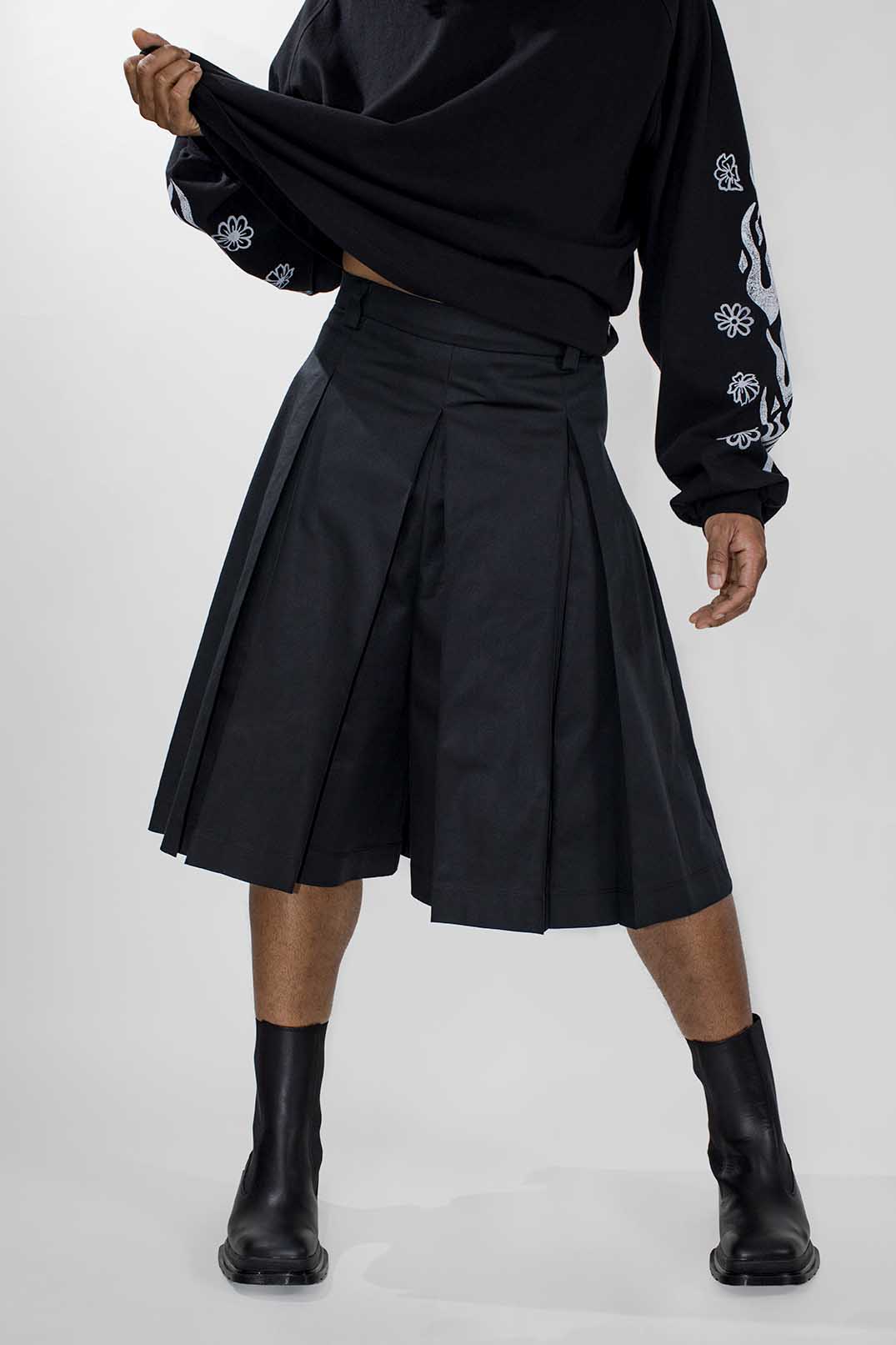 A model wearing a black pleated midi skort, a black long-sleeve top with white graphic prints on the sleeves, and black ankle boots, shown from the waist down against a light gray studio background.