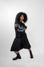 A model with curly black hair poses in a black long-sleeve top with white graphic patterns on the sleeves, a matching black pleated skort, black socks, and black boots against a light gray studio background.