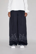 A person wearing very wide-leg black pants with a white flame embroidery along the lower hems, paired with a white top and white platform sneakers, photographed against a light gray studio background.