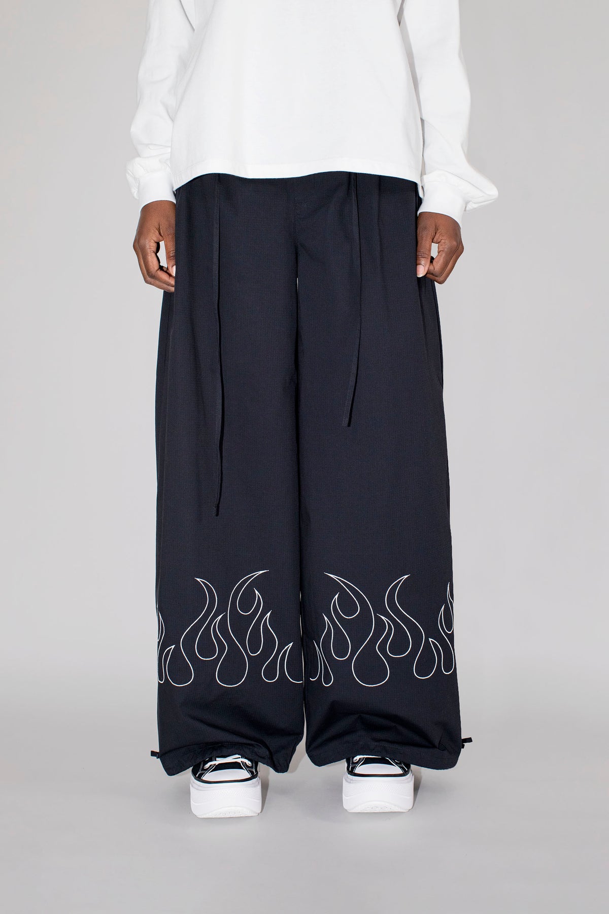 A person wearing very wide-leg black pants with a white flame embroidery along the lower hems, paired with a white top and white platform sneakers, photographed against a light gray studio background.