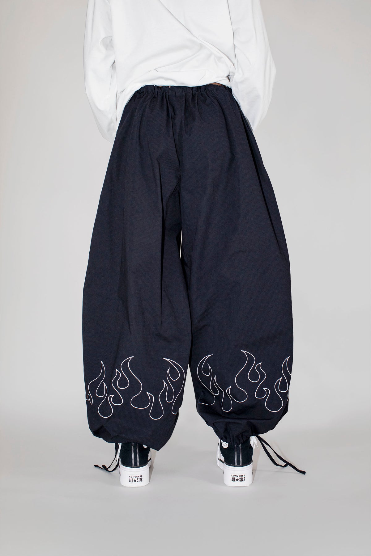 A person shown from the back wearing loose black drawstring pants with white flame embroidery near the hems, a white top, and black-and-white sneakers against a light gray studio background.