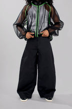 A model wearing a sheer black mesh zip-up jacket with neon green piping over a gray and black patterned top, paired with black wide-leg pants and black sneakers with white soles, set against a plain light gray studio background.