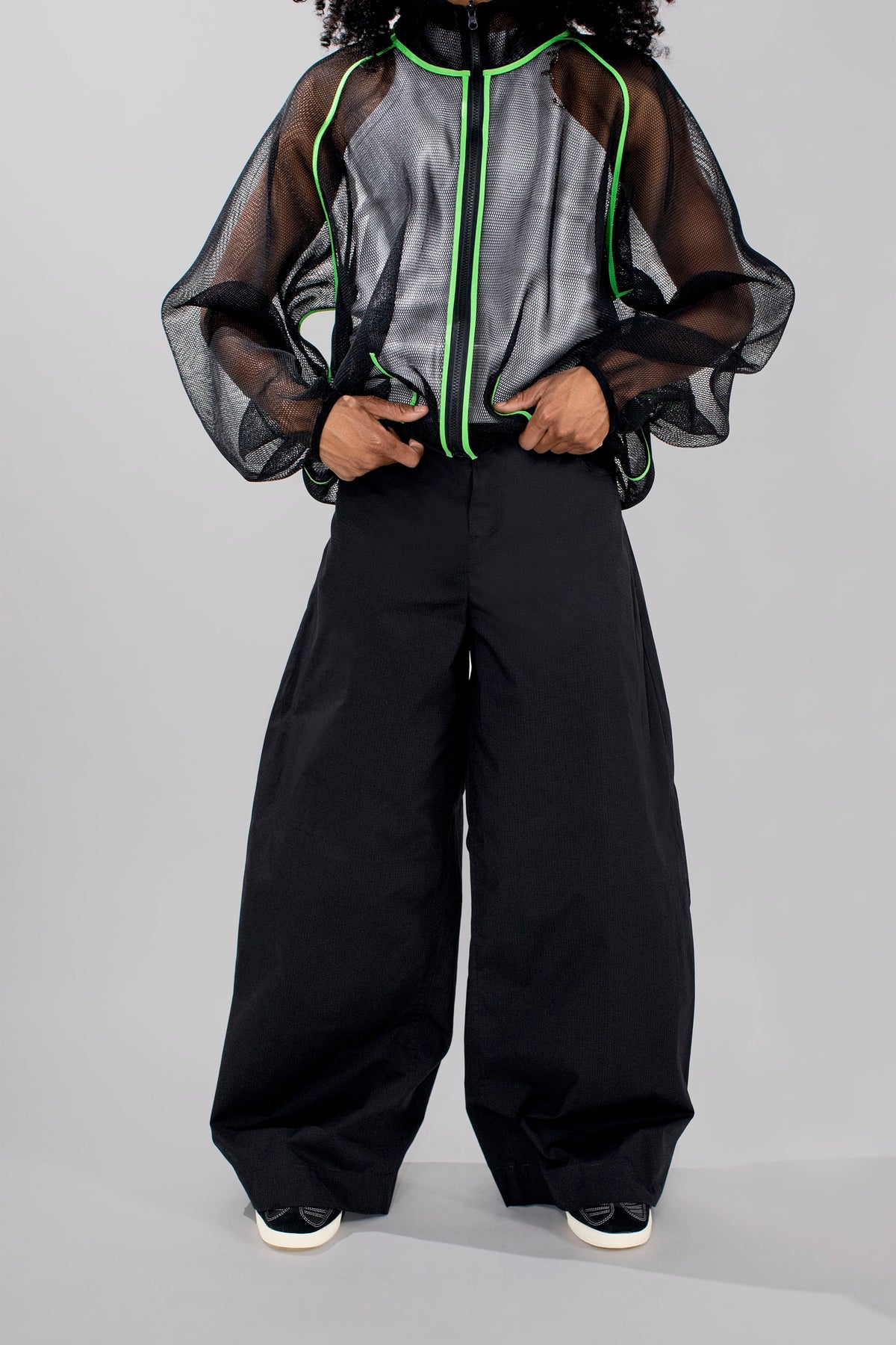 A model wearing a sheer black mesh zip-up jacket with neon green piping over a gray and black patterned top, paired with black wide-leg pants and black sneakers with white soles, set against a plain light gray studio background.