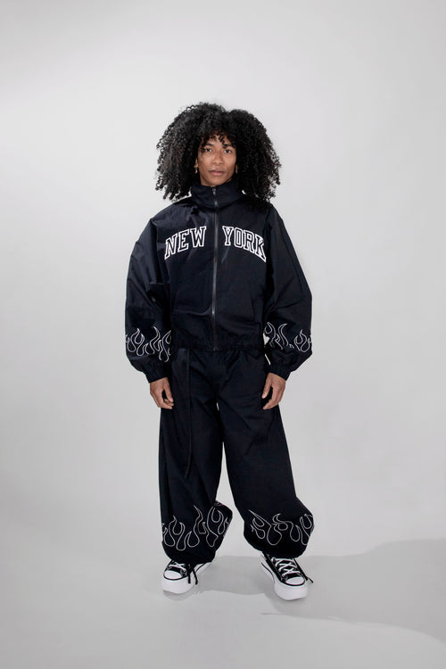 A model with curly black hair wearing a black zip-up jacket and matching wide-leg pants with white embroidery details and 'NEW YORK' text on the chest, paired with black and white sneakers, against a light gray studio background.