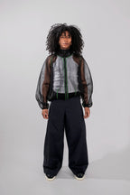 A model with curly black hair stands against a light gray studio background wearing a dark gray and black sheer mesh zip-up jacket over a gray top, paired with loose black trousers and black sneakers.