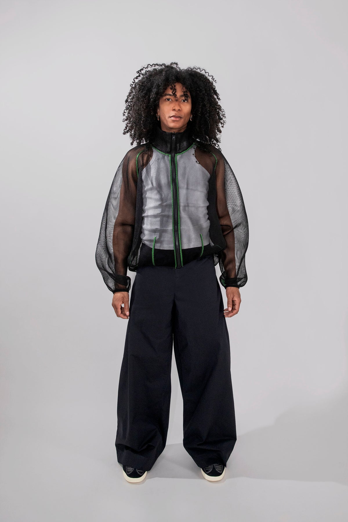 A model with curly black hair stands against a light gray studio background wearing a dark gray and black sheer mesh zip-up jacket over a gray top, paired with loose black trousers and black sneakers.