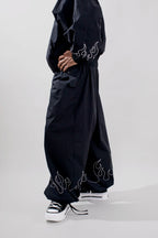 A person wearing loose black wide-leg pants with white flame-like embroidery near the hem, paired with black sneakers, photographed against a light gray studio background.