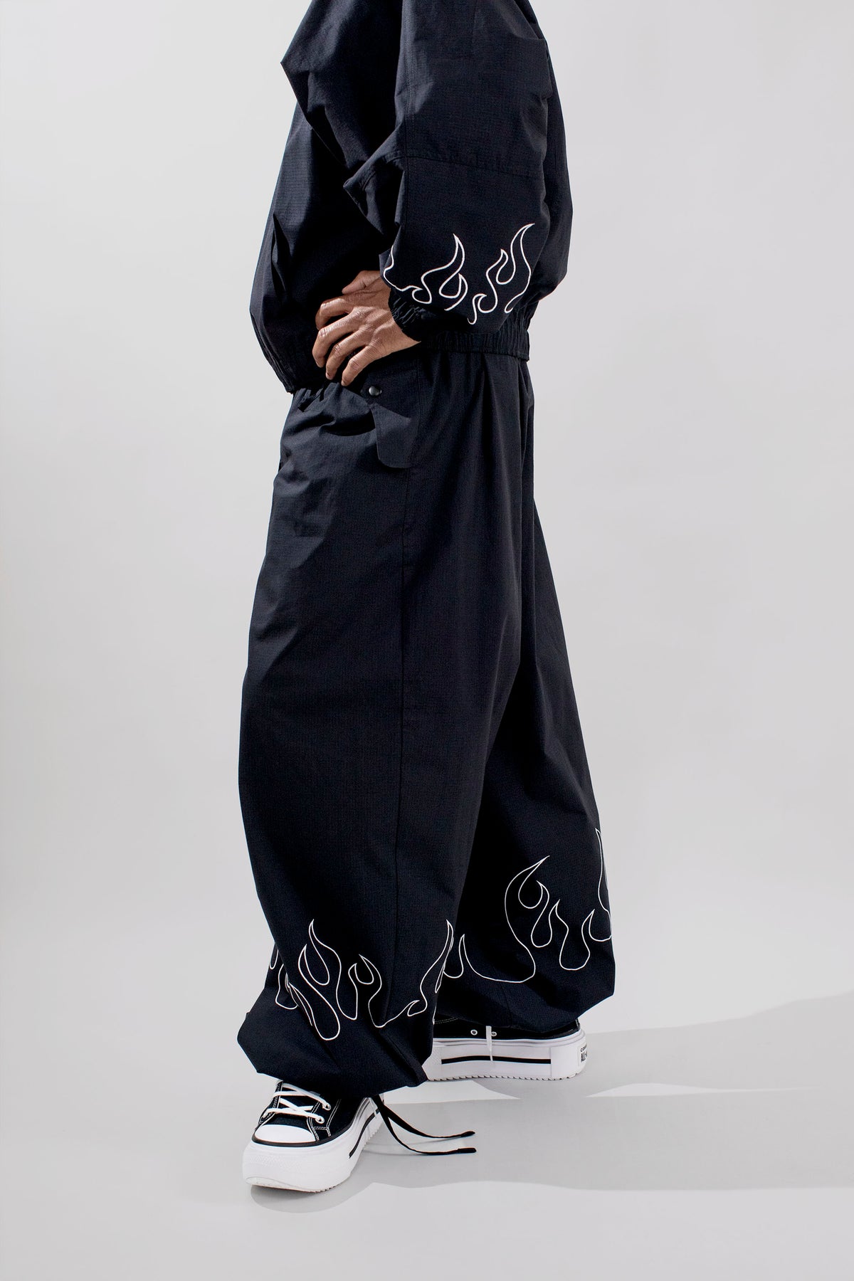 A person wearing loose black wide-leg pants with white flame-like embroidery near the hem, paired with black sneakers, photographed against a light gray studio background.